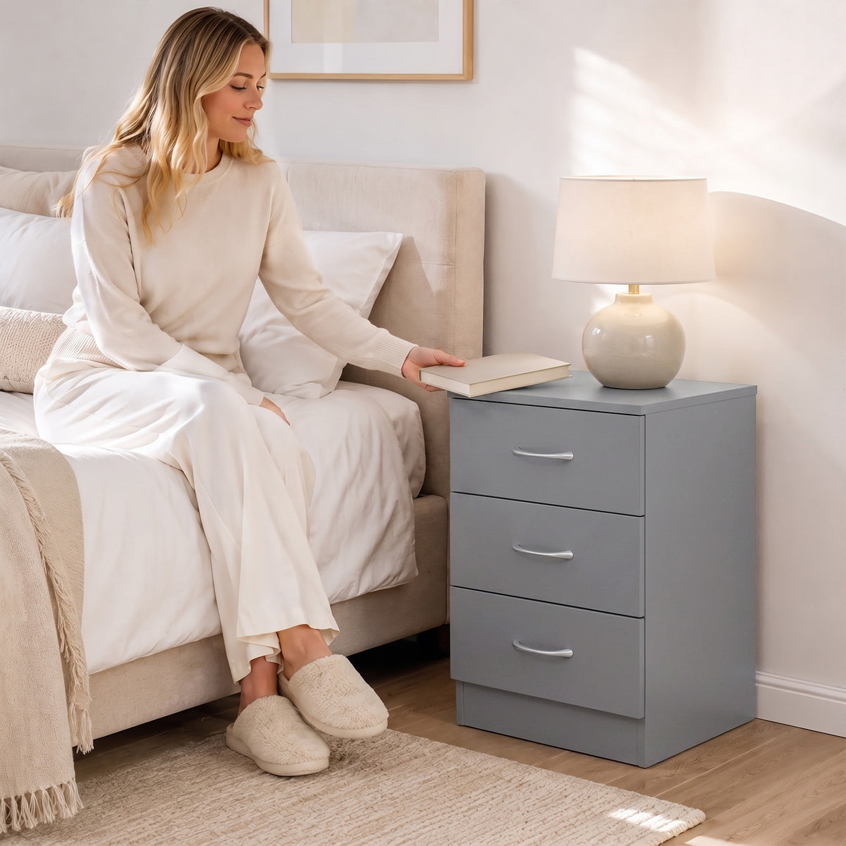 Grey three-drawer bedside cabinet with curved metal handles in a bedroom setting.