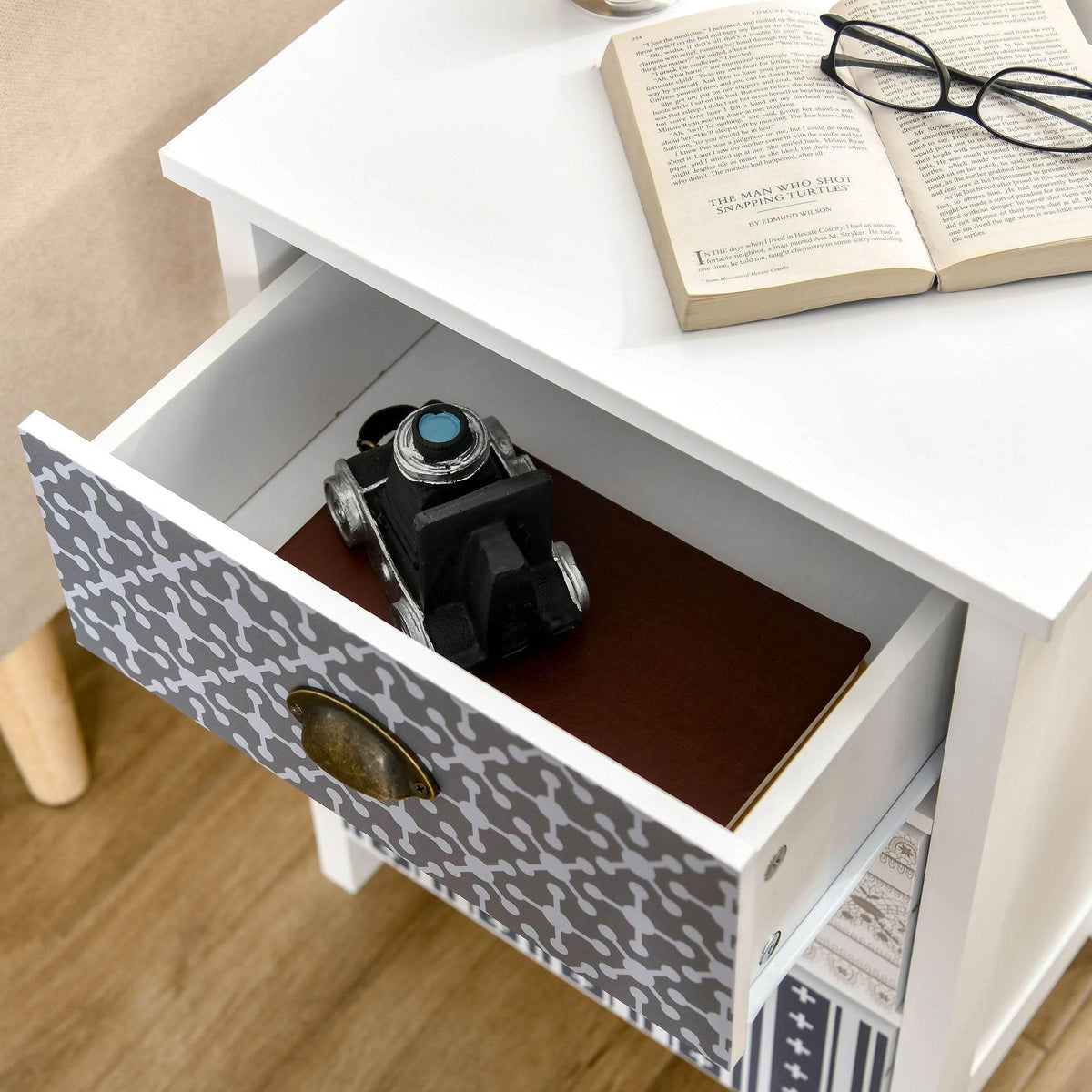 White nightstand with one drawer open showing camera, patterned front and antique brass pull