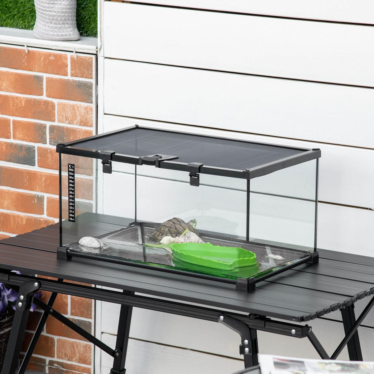 Clear glass reptile terrarium with black frame on stand, inside substrate and green dish