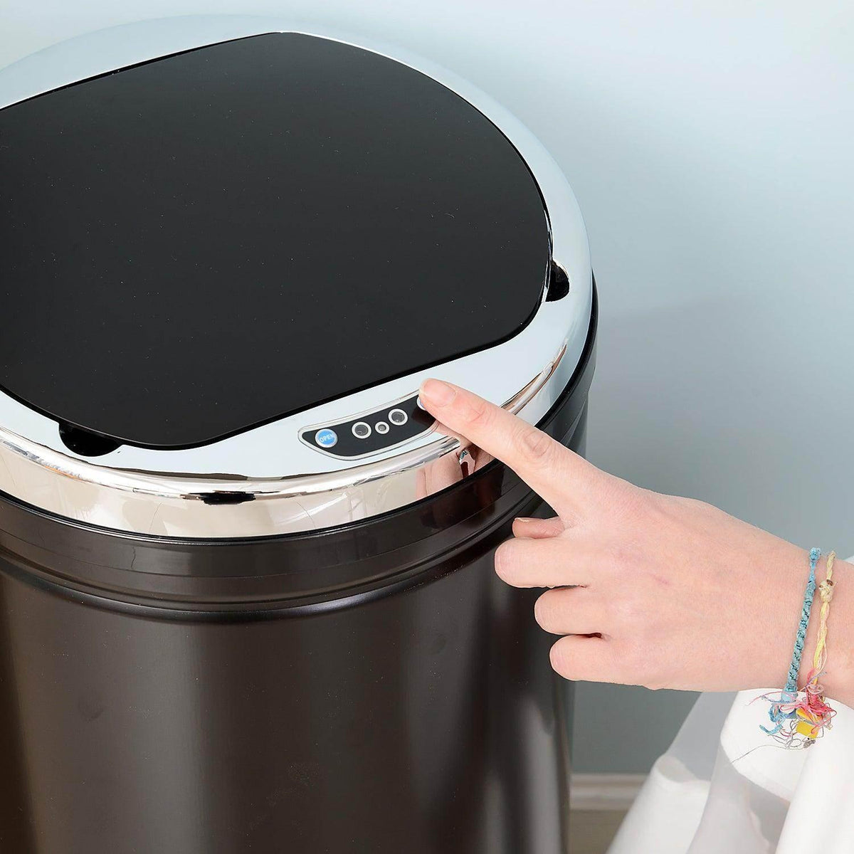 Modern black touchless trash can with chrome lid, hand pressing sensor button for hands-free opening