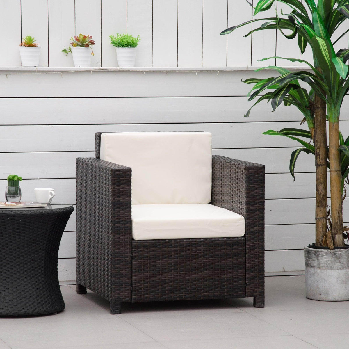 Brown wicker outdoor armchair with cream cushions on patio, beside side table and potted plant