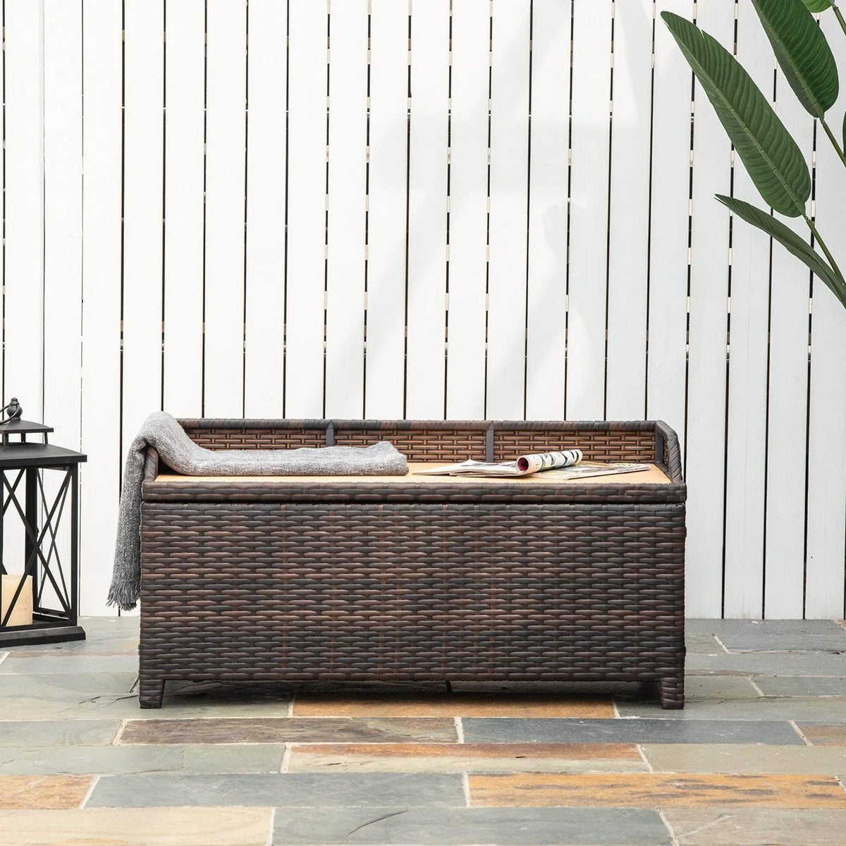 Brown wicker storage bench with gray cushion and folded throw on seat