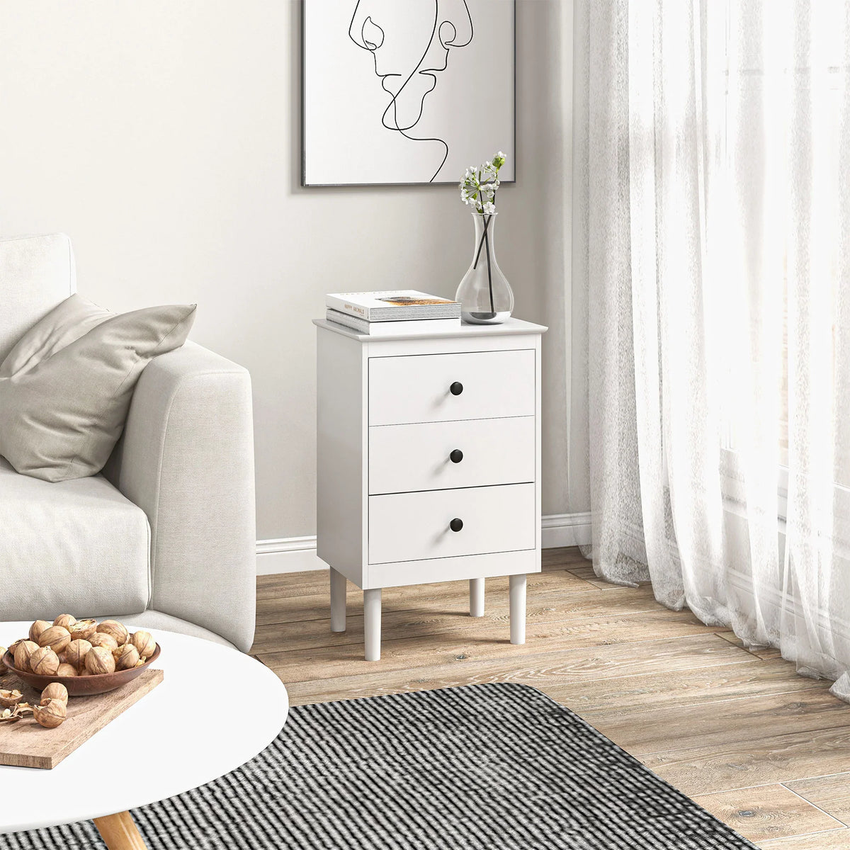 White bedside table with three drawers and round black knobs beside an upholstered sofa