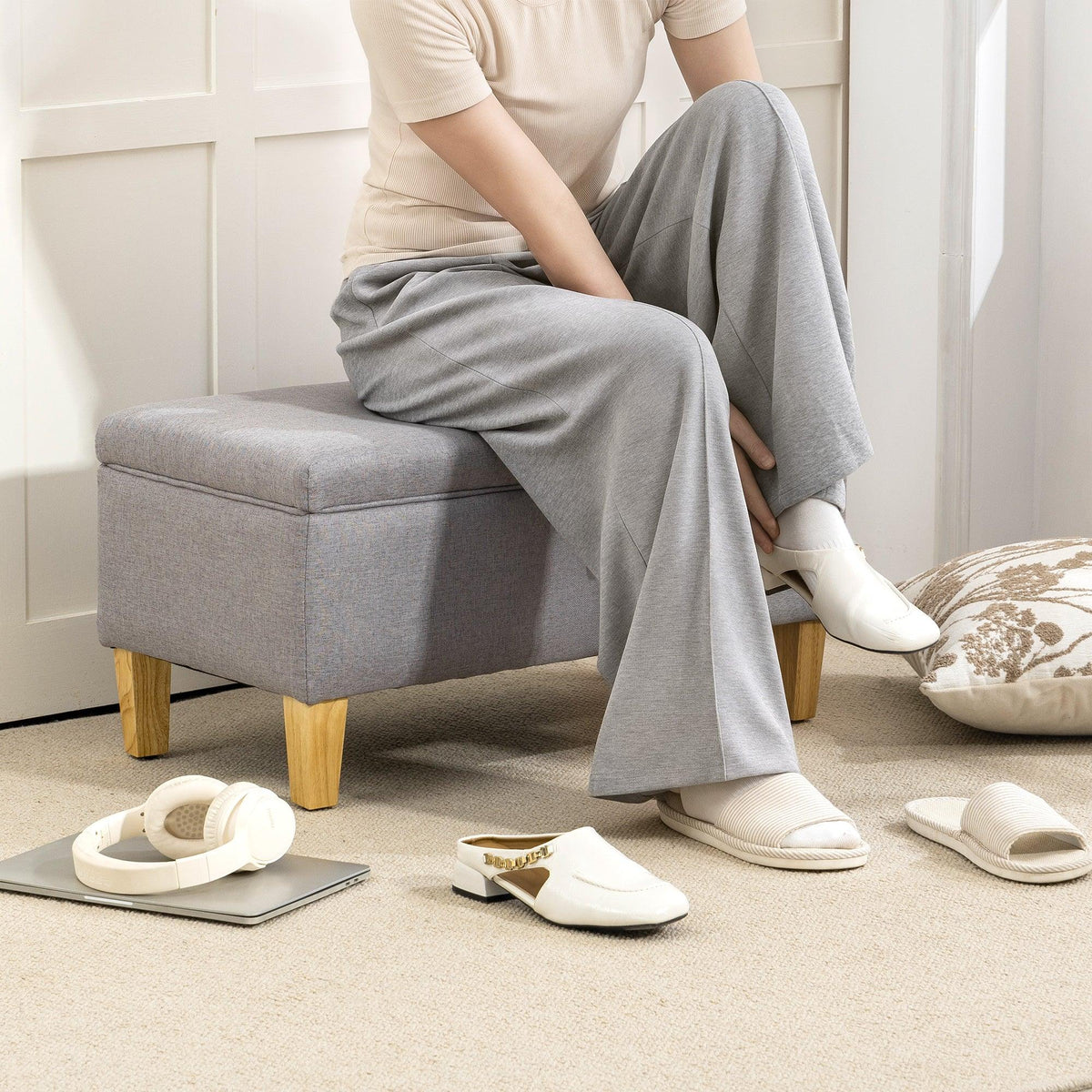 Light grey padded storage bench with wooden legs, person seated with slippers