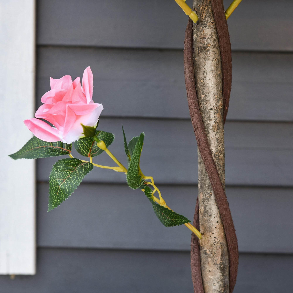 Artificial pink rose blossom on textured brown tree trunk with green leaves in white pot