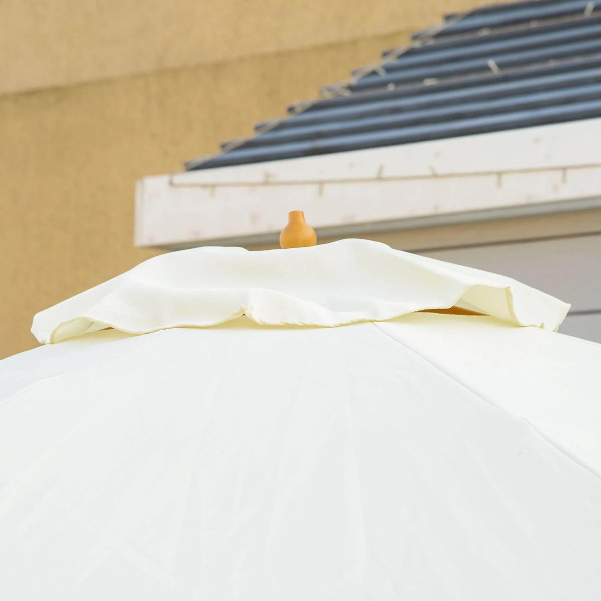 Beige patio umbrella canopy with finial at the top and ruffled edge detail, close-up view.