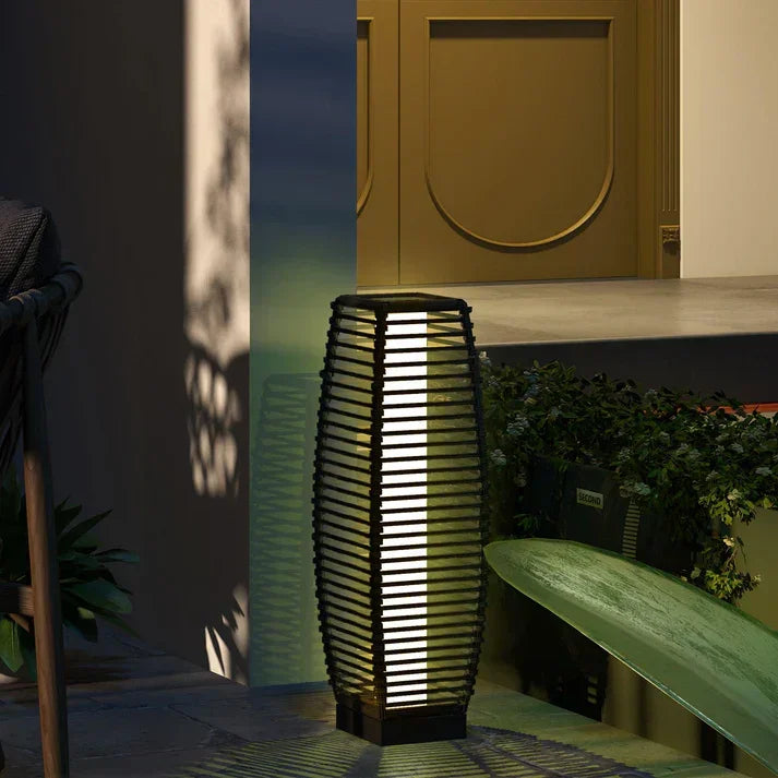Modern outdoor lantern with slatted design, glowing warmly on patio near greenery and gold door