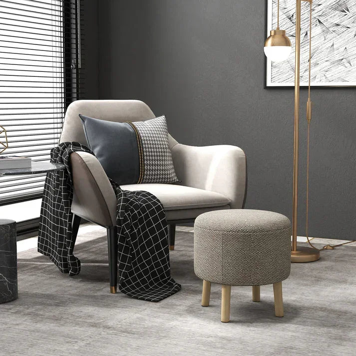 Modern beige armchair with throw pillows, patterned blanket, and round ottoman in cozy living room setting