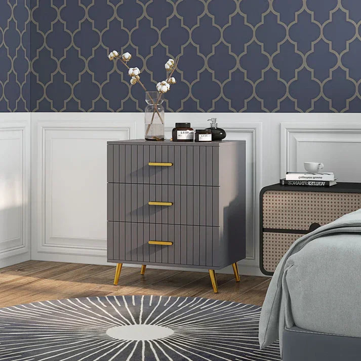 Modern gray 3-drawer nightstand with gold handles, styled in bedroom with patterned wallpaper and rug