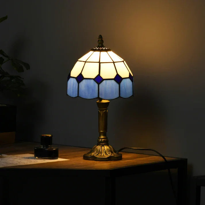 Stained glass table lamp with blue and cream panels, vintage-style base, glowing warmly on wooden desk