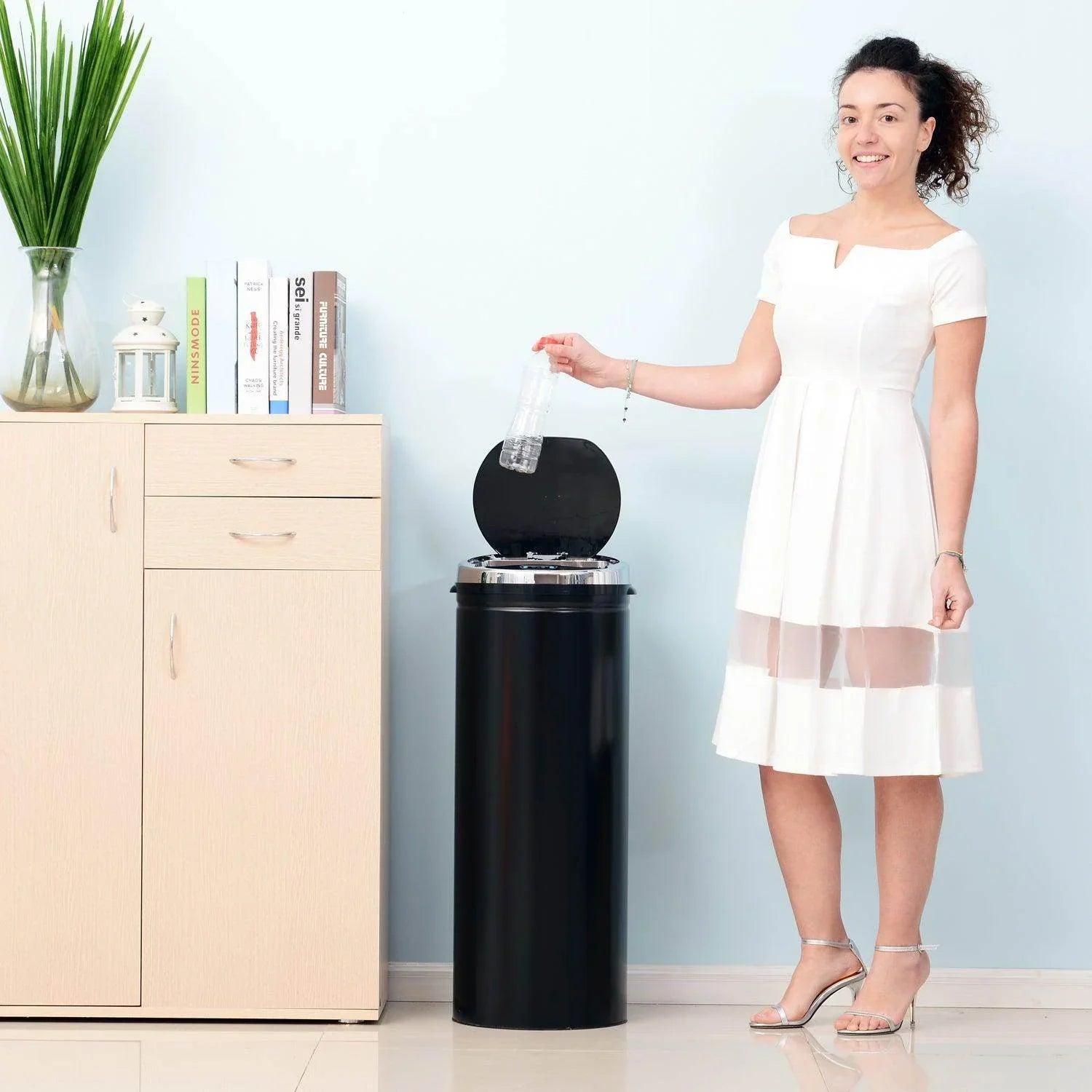 Woman in white dress using black stainless steel sensor trash can with lid in modern home setting
