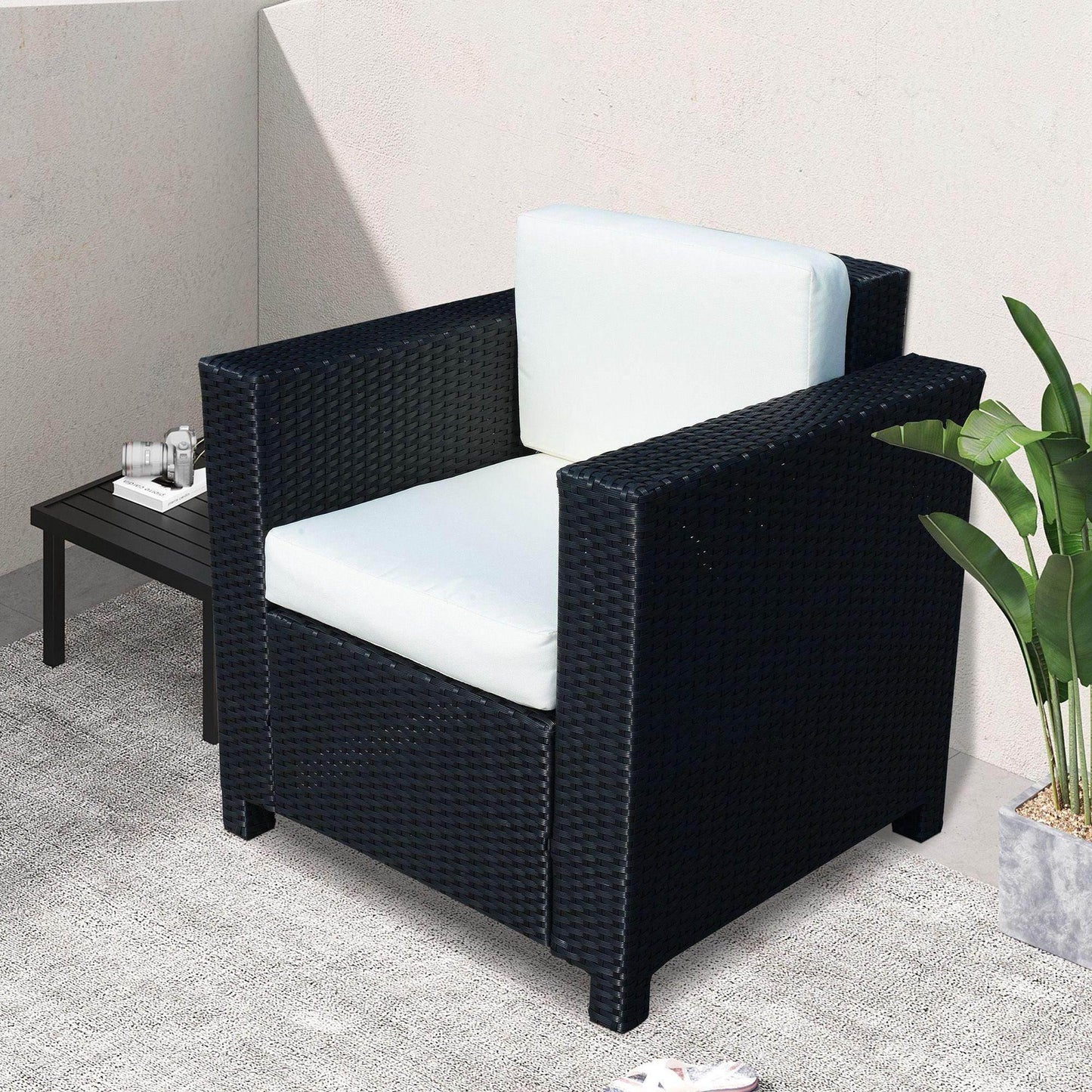 Black rattan single sofa armchair with white cushions on outdoor patio near side table and green plant