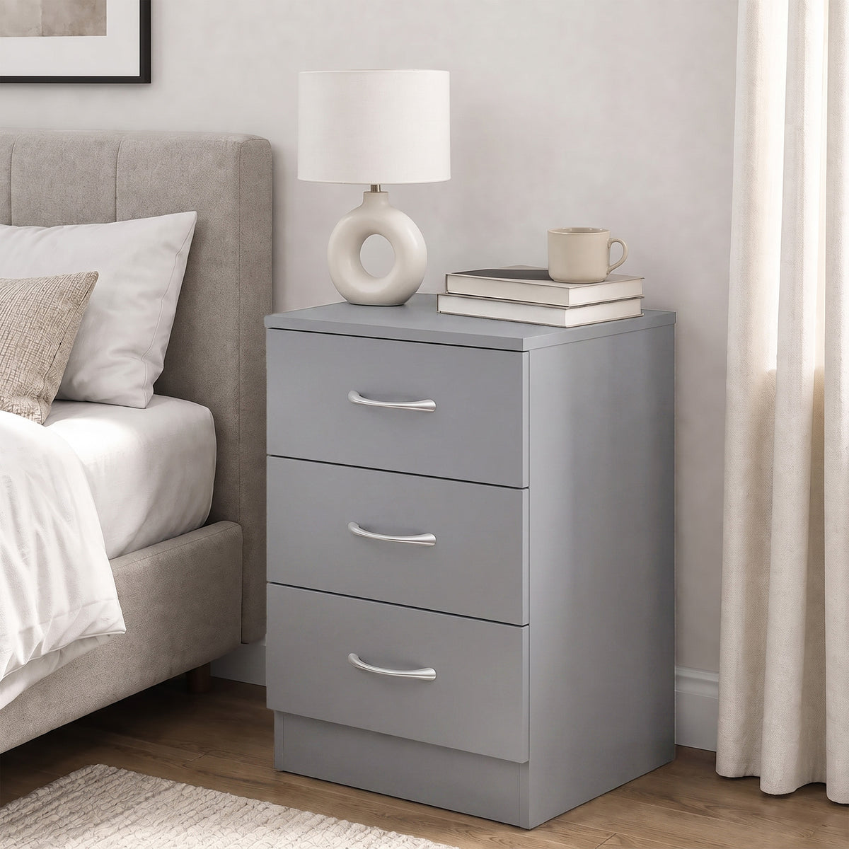 Grey three-drawer nightstand with silver curved handles beside a bed.